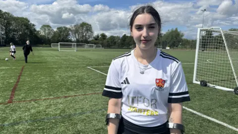 Zoie O'Brien/BBC A head and shoulders image of a young woman wearing a white and black football shirt. She is standing on a football pitch with a goal behind her. She is smiling and looking into the camera.