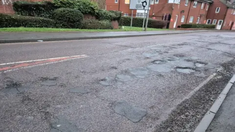 Swindon Borough Council A large cluster of poorly patched potholes on one side of the road, with a block of residential houses and flats visible in the background. 