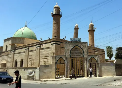 EPA people walk in front of the al-Noori al-Cabeer mosque in Mosul. The mosque green dome is visible behind the main gate, and the leaning Minaret strikes a blue, cloudless sky in the background.