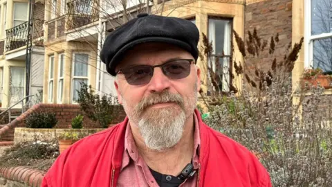 Resident of Hotwells Steve Page, wearing a black cap, sunglasses and a red jacket. He has a grey beard. 