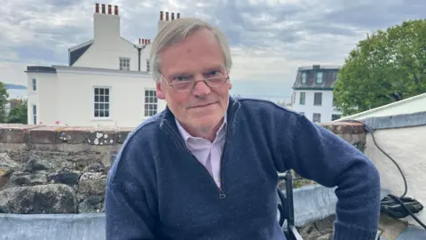Simon Thornton - A man with grey hair, and half moon spectacles looking at the camera. He is sitting down on a chair on the Old Government House balcony and leaning on his right knee with his right arm. He is wearing a blue quarter zip on top of a purple button up shirt. 