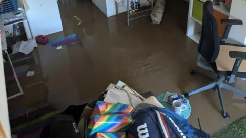 Ben Perham A room in a house in Ilminster in Somerset that is submerged in brown flood water. Various belongings have been piled into a box to be safe from the water and furniture can be seen standing in it