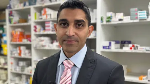 Sanjeev Panesar stands in one of his shops in Sheldon, Brimigham. He is wearing a pale blue shirt, pink striped tie and dark jacket.