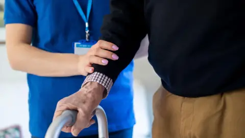 A carer wearing a blue tunic and a lanyard around her neck helps an old man. Only their torsos are visible. The man has one hand on a zimmer frame and is wearing a checked shirt under a black jumper and beige trousers. The carer has pink nails and is resting a hand on the man's lower arm.