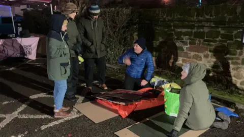 Five people are sitting or standing getting ready to take part in the sleep out. Cardboard and sleeping blankets are on the ground and everyone is wearing a coat and headwear.