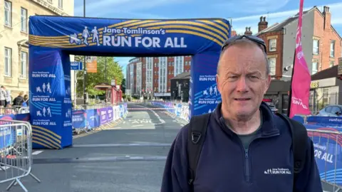 A man in his sixties in a navy fleece with "Jane Tomlinson's Run For All" written on it in white. He looks at the camera. Behind him the race finish line arch has the same branding on it.