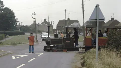 Chris Grimes A person standing a road, holding a red flag, with a train going past, and cars and houses in the distance. 
