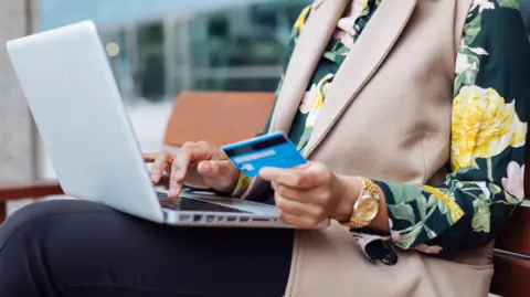 A woman in a flowery blouse holds her bank card up to make a payment on a laptop