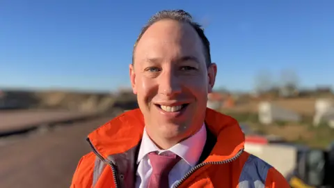 Alex Dunlop/BBC Tom Farley is standing outside near a section of the A47 where works are being carried out. He is looking directly at the camera and smiling. He is wearing an orange fluorescent hi-vis jacket over the top of a white shirt and red tie. The sky is blue in the background behind him.
