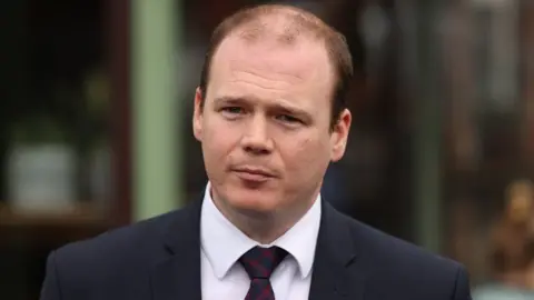 PA Media Gordon Lyons has short brown/red hair which is balding on top. He is wearing a dark suit jacket, white shirt and red and black tie.