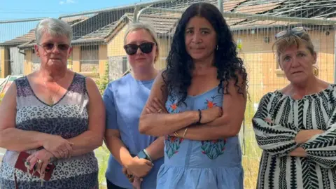 Four women stand outside a barrier entrance to the building with their arms folded looking unhappy about its derelict state.