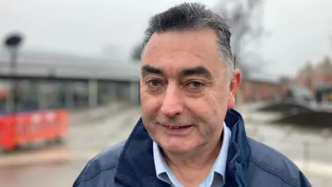 A man with short black hair peppered with grey grins at the camera. We can see plastic orange barriers at a railway station in the background