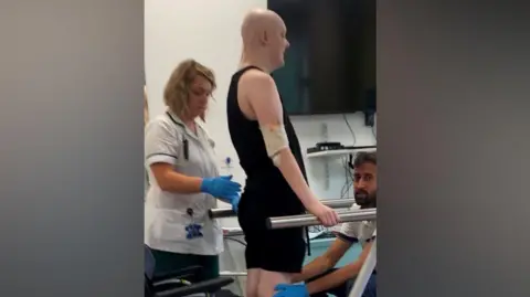 Supplied A young man with black trousers and a black vest being supported while standing up in a hospital room by two nurses.