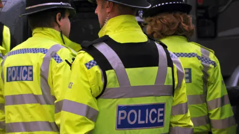 A stock image showing the back of three police officers. They are wearing hi-vis yellow flourescent jackets with the word Police across the back. They are also wearing helmets. 