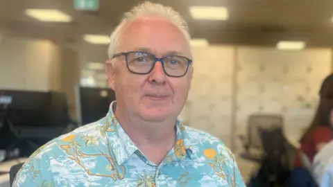 Dave Rigby, senior forecaster at Jersey Met is smiling at the camera. He is wearing glasses and a tropical-print shirt and is sat in an office. There are two colleagues working at computers slightly blurred in the background.