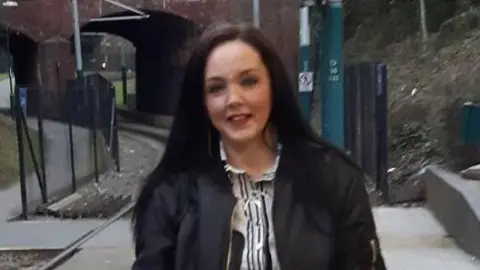 Lincolnshire Police Ellen Marshall with dark brown long hair and wearing a black bomber jacket over a monochrome striped dress. She is standing on a train platform with the train tracks visible on the left