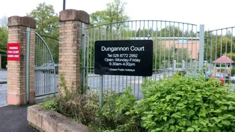 railings outside dungannon court car park show a sing with the courts opening hours. parked cars can be seen through the metal bars