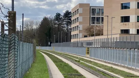 Jozef Hall/BBC A photograph of a section of the Cambridge busway. Grass is growing on either sides of the concrete of grey concrete bordered by green grass. A line of grey metal fencing runs along both sides of the busway. It is a blue sky day but with some white clouds.