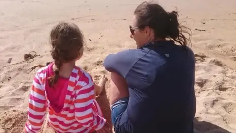 Nicola Allen Nicola and her daughter sitting on a beach facing away from the camera. Nicola has her dark hair tied back. Her face is turned to the side and she is wearing sunglasses and a dark blue top. Her daughter, on the left, has brown hair in a plait down her back. She is wearing a pink, red and white striped hoodie.