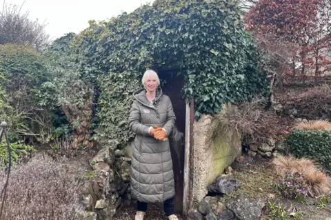 A woman with short white hair smiles as she stands in front of the doorway to a large concrete air raid shelter which is almost completely covered by green ivy. She is wearing a long green coat.