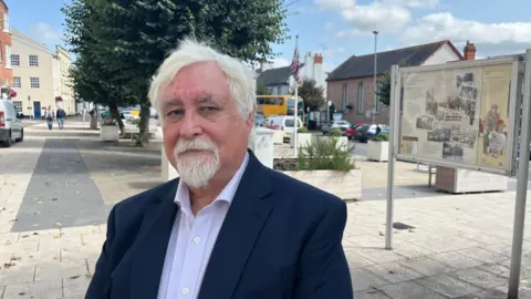 Councillor Steve Keable, standing near the war memorial in Cullompton. He has white hair, a white beard and moustache, and is wearing a light blue shirt and navy blazer.