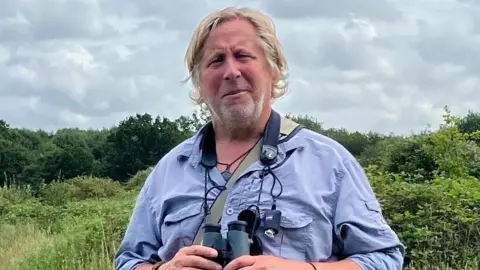 Supplied A man wearing a blue shirt and holding a pair of binoculars which are hanging around his neck. He is standing in a woodland with trees and bushes in the background.