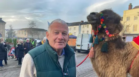 Alasdair Elliott, wearing a cream jumper with a green gilet stood next to Baxter the camel with colourful tack