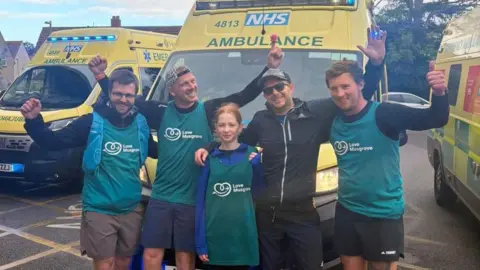 A group of people are standing next to each other raising their arms. There are yellow ambulance vans behind them.