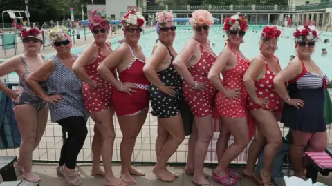 A group of nine women standing in a line with their hands on their hips. They are all standing towards the right with their heads turned towards the camera and are smiling. A majority of them are wearing polka dot swim suits and have sunglasses and floral swimming caps on.