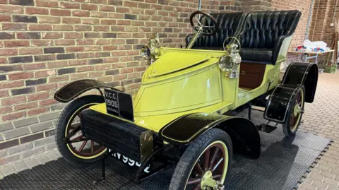 Stockwood Discovery Centre A vintage car with two raised leather seats and an exposed cabin. It is yellow and the wheels have yellow rims. A plate on the front reads: V.C.C 1905 Vauxhall. The car is parked in a building on rubber matting by a brown wall.