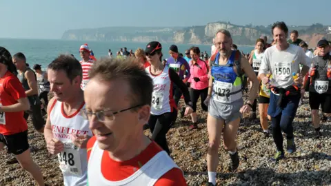 A large group of runners jog along Seaton beach in Devon as they take part in a race called The Grizzly. Cliffs and the sea can be seen in the background.