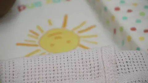 A baby changing mat with a cartoon sunshine at the bottom. It is yellow, and laid on top is a knitted blanket.