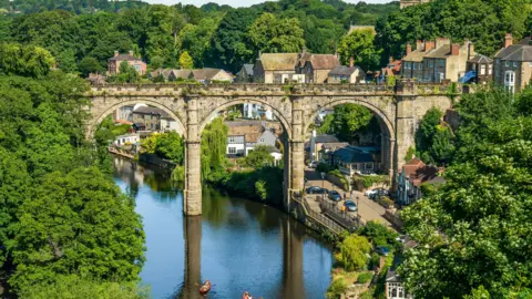 Knaresborough Viaduct, pictured from a short distance away. It stands above a river. Trees surround it. The viaduct has three arches and there are houses in the background.