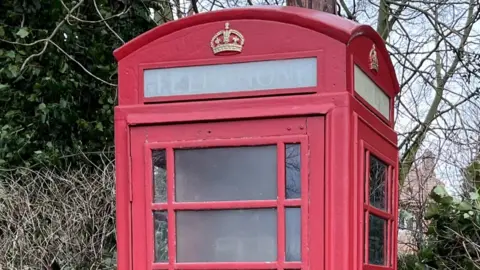 A traditional red phone box stands by a wooden fence with trees behind it.