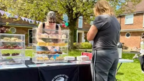 Sarah Lewis A woman with brown hair is preparing cupcakes behind a market stall. There is bunting in the background and balloons, strung between trees. A woman is waiting for cupcakes at the stall.