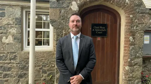 White man in a grey suit standing in front of a brown wooden door that has a black sign that says Chief Pleas & Seneschal's Court. There is a white flag pole next to him.