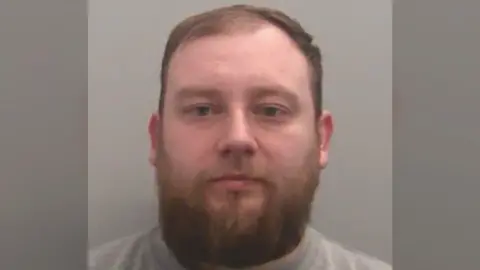 NCA Sam Elphick's mug shot. A man with short brown hair a thick, dark beard.