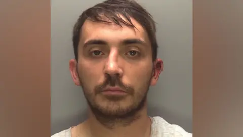A police custody image of a man with dark hair, He is wearing a white top and he has a beard. He is looking directly at the camera.