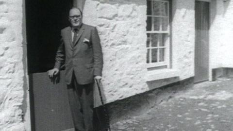 Black and white image of James Boyce leaning on a half door of a cottage.  The rest of the building is whitewashed and there is a windowframe behind and another door behind on the same wall.