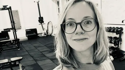 Hannah Buckley Hannah smiles in a selfie in a gym. She has shoulder-length blonde hair and wears dark-rimmed glasses. There is exercise equipment, including weights and benches, visible behind her. 
