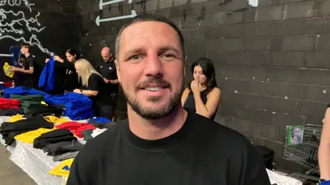 BBC Tommy Coyle pictured at his school uniform giveaway during 2024. He has short black hair and a black beard and is wearing a black T-shirt. Behind him is a long table with various multi-coloured items of school uniform laid out. Five people can be seen behind the table who are working on handing out the items of clothing. 