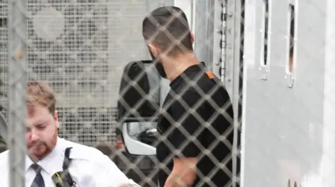 Eddie Mitchell A man in a dark T‑shirt is near a van, photographed through metal fencing, with a security officer in the foreground.