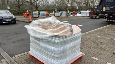 A stack of bottled water sits on a pallet in a car park. In the background there are people in high vis jackets offloading water bottles from a lorry