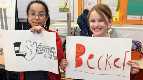 Two girls smile as they hold up two small posters. One has the name Lydia on it, written in black ink with alternating letters filled in with black and white stripes. The second has the surname Becker written in red ink, with the enclosed sections of the capital "B" and the "e"s entirely filled in.