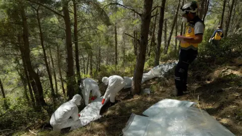 A man wearing a white helmet stands on a slope in a forest. Below him, three people in personal protective clothing attend to a body bag 