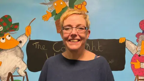 A woman with fair hair and a blue top with a cartoon of the Wombles behind her.