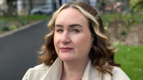 BBC A close-up image of Fiona Ferris, who has dark brown, shoulder-length hair and a blonde fringe. She is looking at the camera with a neutral expression on her face. She is wearing a light coloured coat and is sitting on a bench in a park, but the background is blurred.