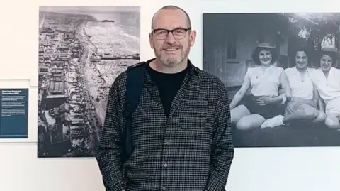 Steven Howells, who is wearing a shirt with a black T-shirt underneath and is smiling at the camera, is standing in front of a white wall with black and white photographs take by his grandad. The picture on the left is an aerial photograph of the coast and the image on the right is on three women sitting on the grass in front of a house. 