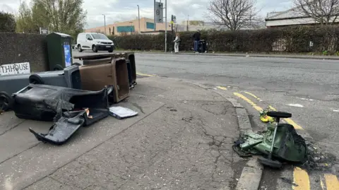 Melted containers on the side of Captain's Road. A green container is at the curb on double yellow lines and has melted badly. It has two wheels joined together and one of them is suspended in the air. A black container is partially melted on the pavement. It is next to another black container and two brown containers that are not affected. 