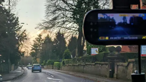 A car with dashcam in operation. Recording info can be seen at the bottom of the rearview mirror. There are cars on the road in front. The car is driving down a  road with houses, walls, hedges and trees.